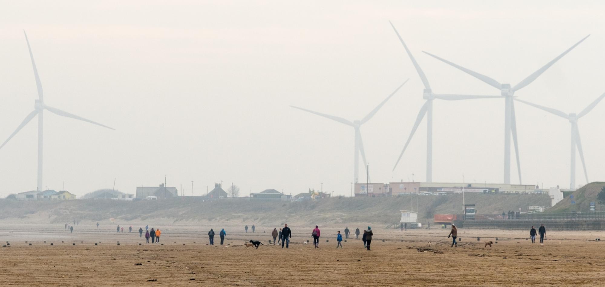 A photo of a beach, in the distance several people are walking along it. In the far distance, the outlines of wind turbines can be seen.