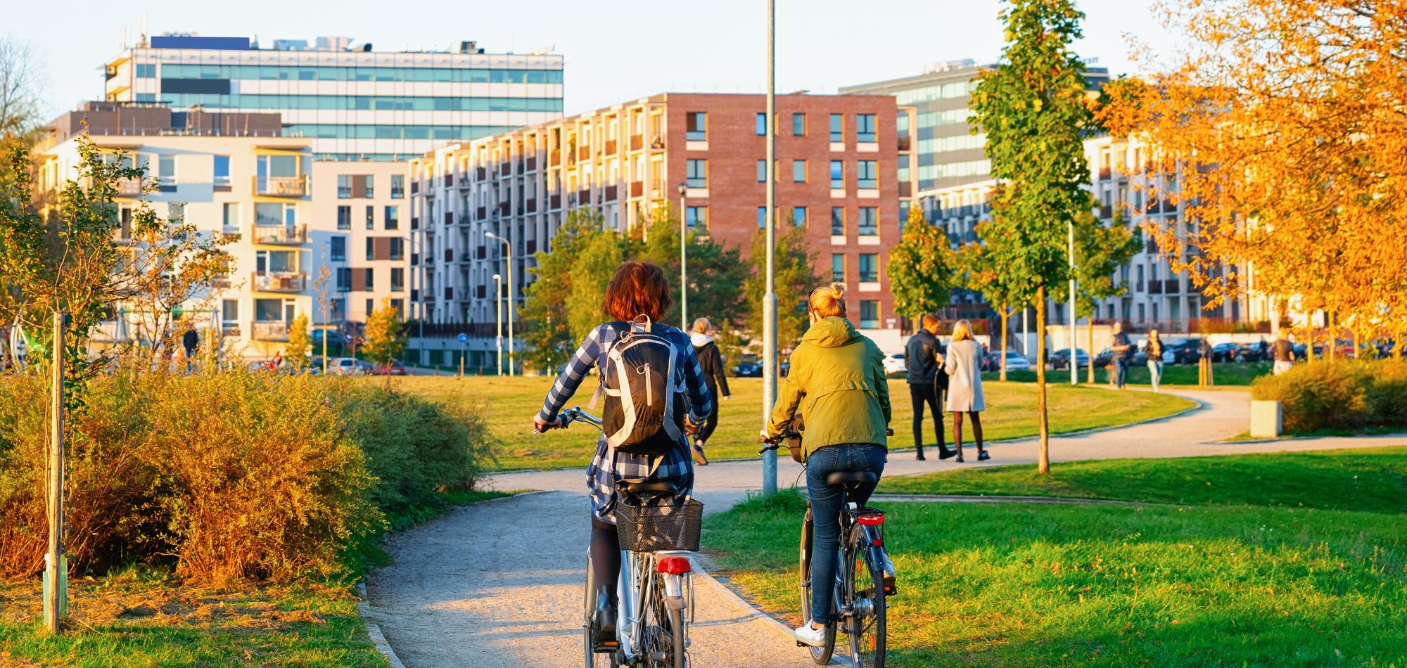 Two cyclists riding on a path in a park with modern buildings and trees in the background during sunset.