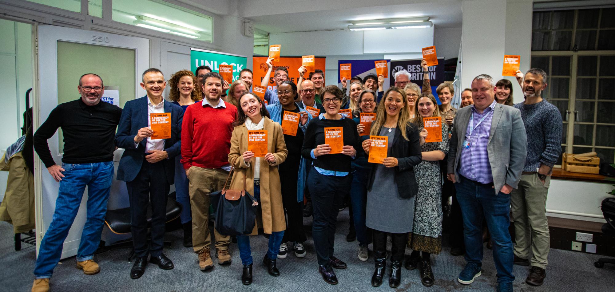 A group of people holding orange flyers that read 'it's time for a democracy fit for the 21st century'