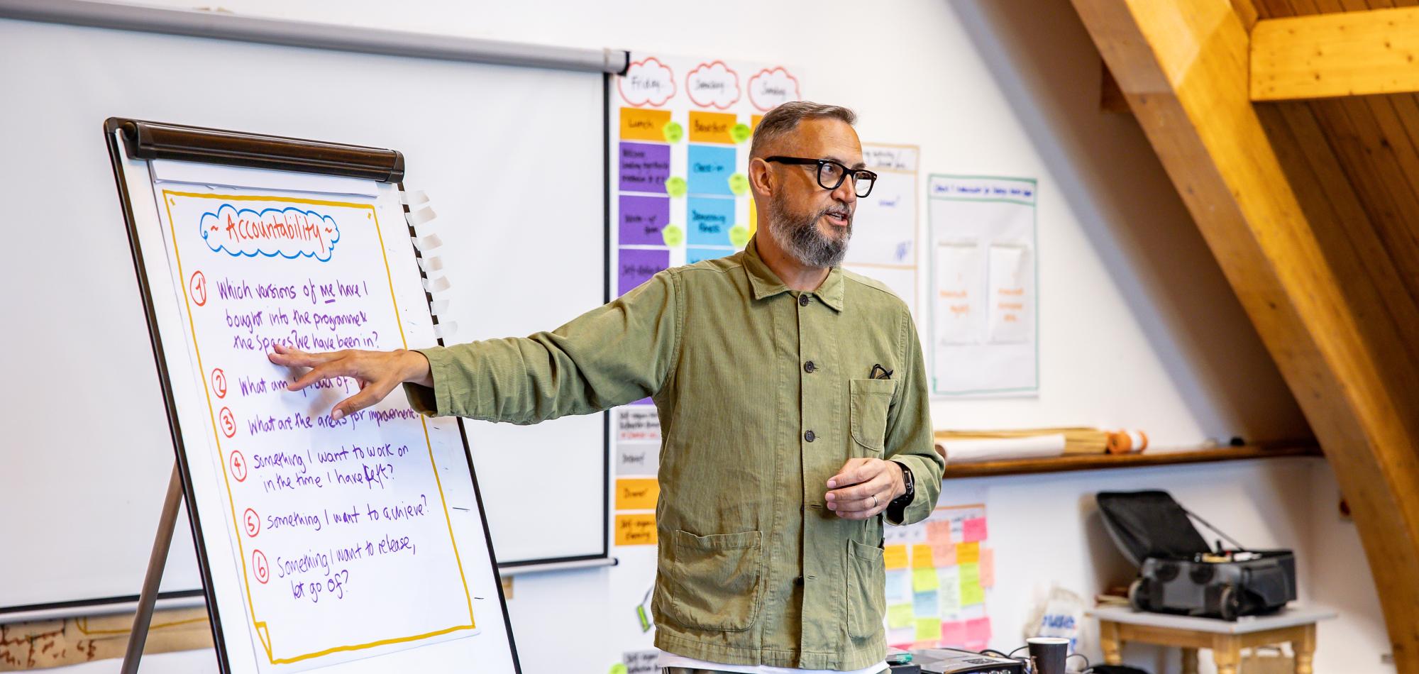 A man wearing a green shirt, pointing to a flip chart on a board.