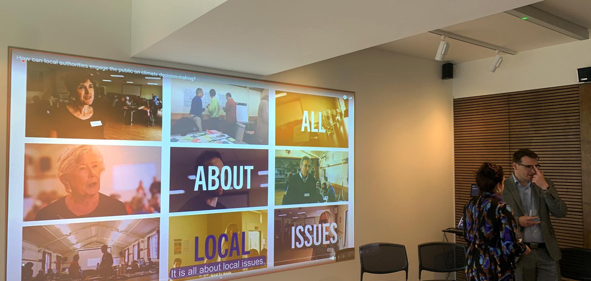 Image of a projector screen showcasing a video with two people standing by.