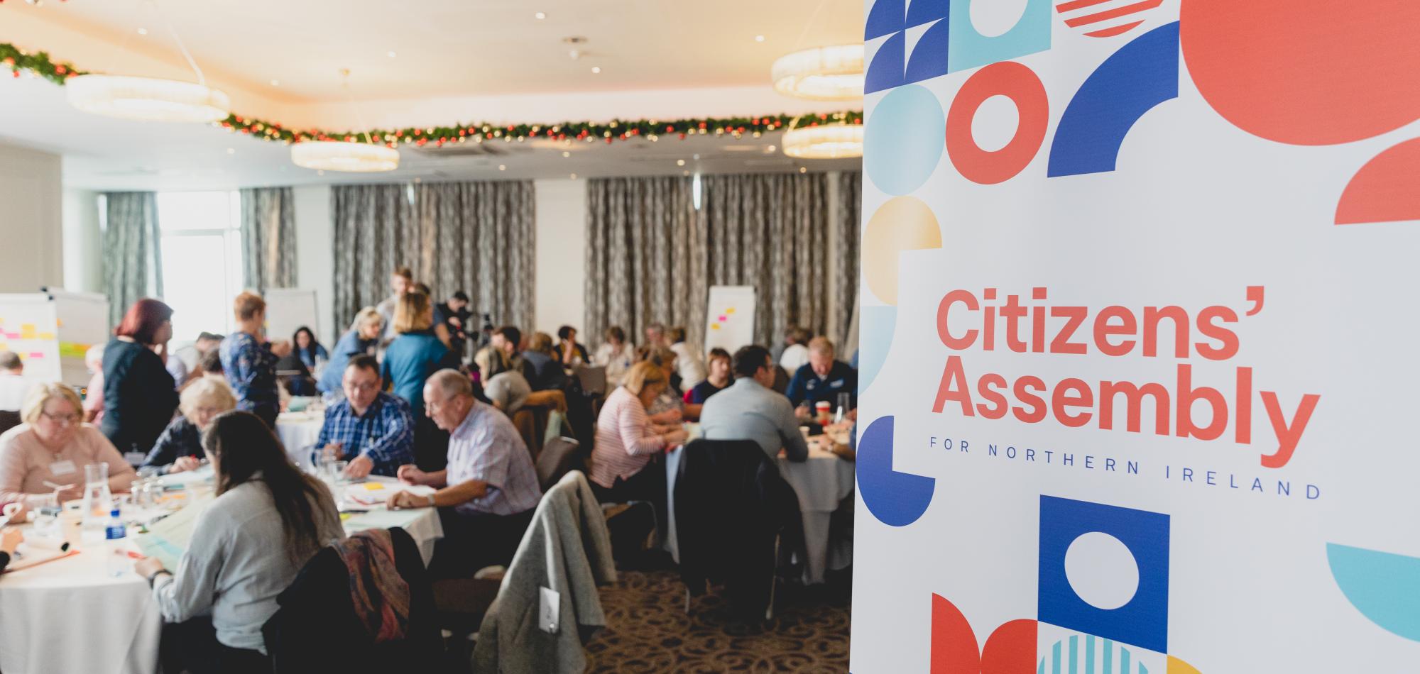 Image of the Northern Ireland Citizens' Assembly at work, in the foreground is a banner that reads 'Citizens' Assembly for Norther Ireland'