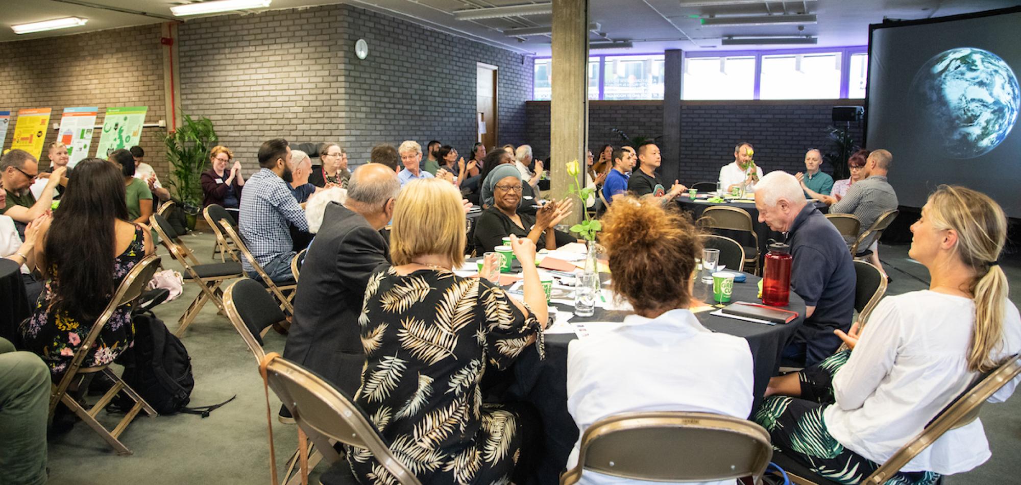 A room full of citizens sitting around round tables, all are smiling and applauding. In the background, an image of the earth is on a large screen. 