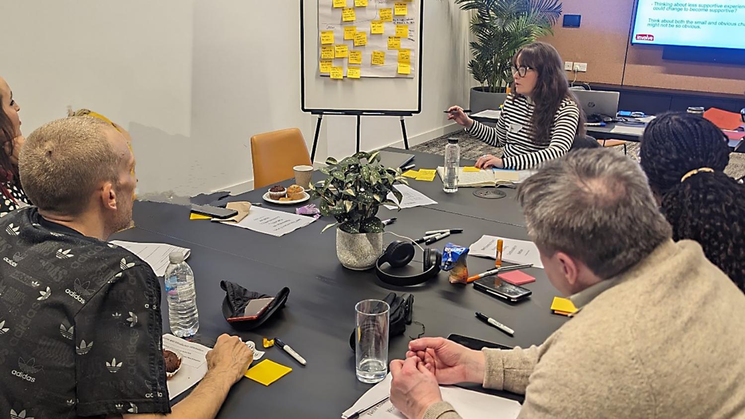 Picture of people in a room talking about ideas. A presentation is in the background and a flipchart with sticky notes.
