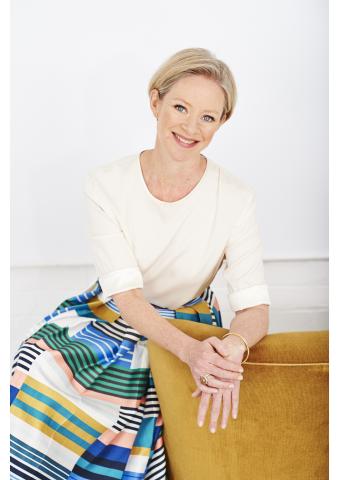 A smiling person with short blond hair, wearing a cream top and colorful striped skirt, leans casually on a mustard chair, exuding warmth and confidence.